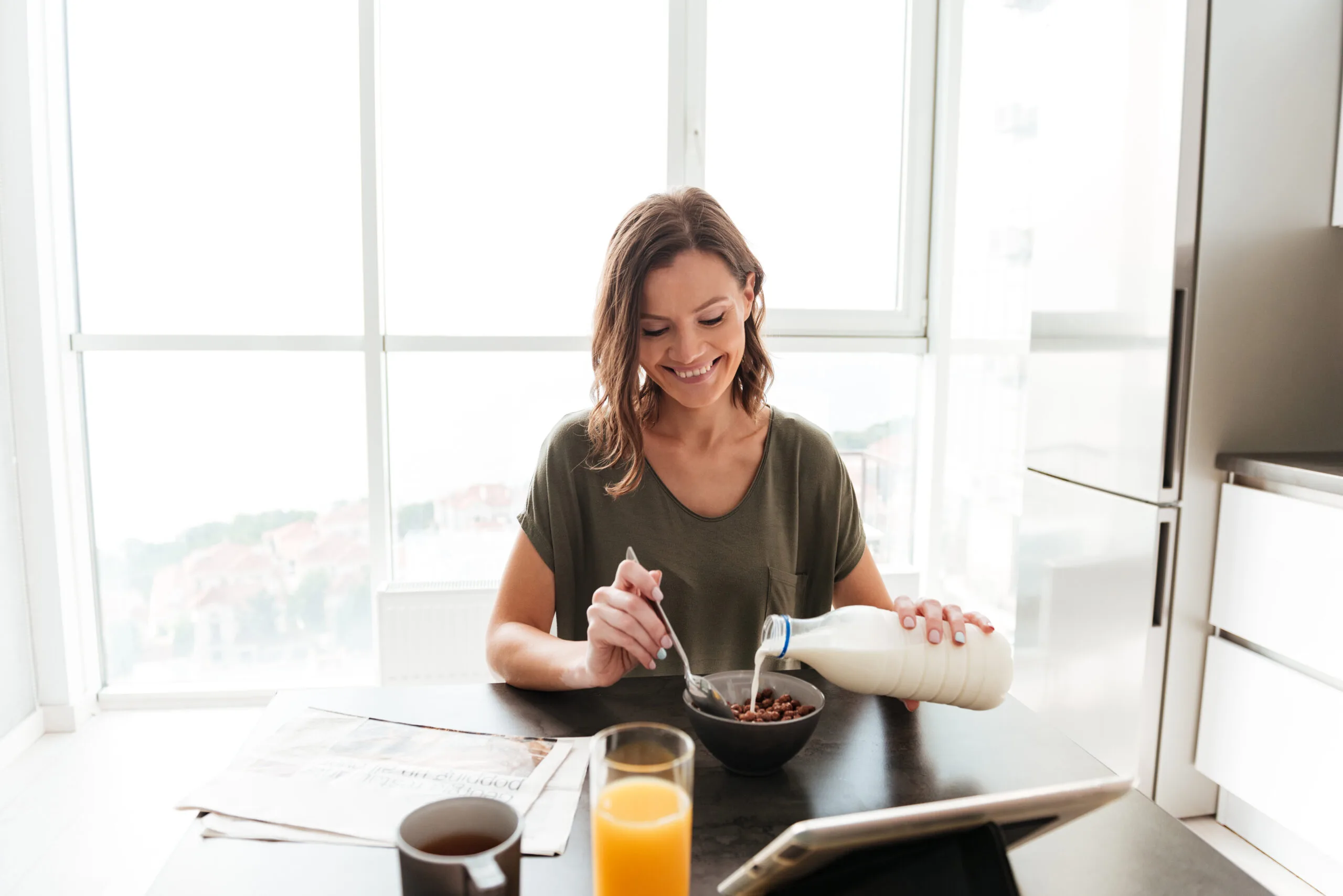 Pleased casual woman eating by the table on kitchen and looking at the tablet computer